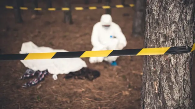 Ormanlık bir alanda, üzerindeki beyaz tulum ile yerde eğilmiş çalışan bir olay yeri inceleme personeli. 