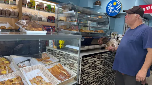 Walter Díaz frente a la vidriera de la panadería La ROU. Viste una camiseta azul manga corta, un pantalón gris y un gorro de visera marrón.