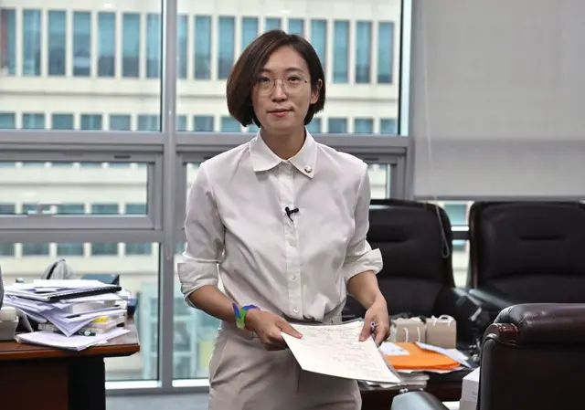 Jang Hye-Yeong dans son bureau en hauteur, vêtue d'une chemise blanche, d'une montre aux couleurs de l'arc-en-ciel et tenant une pile de documents - elle a les cheveux courts et sourit.