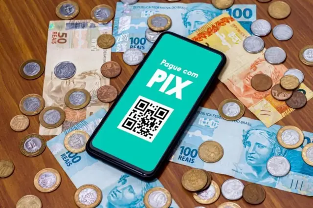 Foto ilustrativa de celular com notas e moedas ao redor. Há uma mensagem escrita 'Pague com Pix' na tela do celular