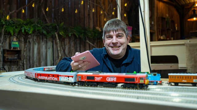 Un hombre está junto a una vía de tren en miniatura que incluye un tren en miniatura principalmente rojo y blanco.