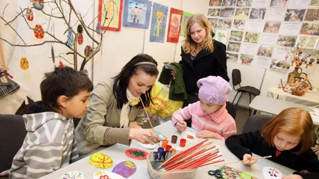 Вчителька малювання разом з кількома дітьми малює паски у кімнаті, прикрашеній дитячими малюнками та поробками.