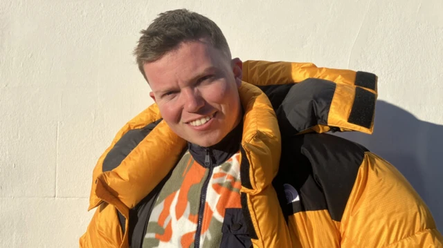 Un hombre con un abrigo acolchado amarillo brillante sonríe a la cámara. Tiene el pelo corto y castaño y lleva un forro polar estampado debajo. Está de pie frente a una pared color crema.