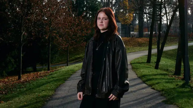 Середній план Камелії. Дівчина стоїть на асфальтованій доріжці в парку. Надворі осінь. Вона вдягнена в чорні широкі штани, чорну шкірянку та чорний светр. Камелія дивиться в об'єктив камери