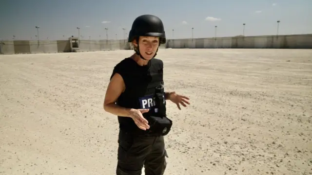 Lucy Williamson pictured at a new aid site which will be run by the GHF. She stands outside on pale brown, stony ground, with a grey fence rising on two sides behind her. She is wearing a helmet and a protective vest with 'press' written on it.