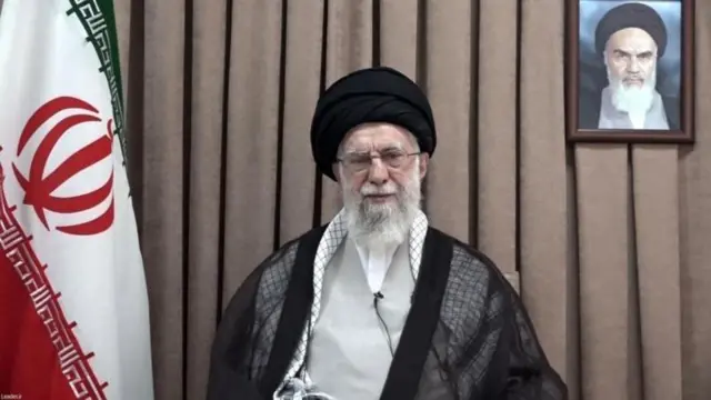 Ayatollah Ali Khamenei yicaye, yambaye imyenda y'umukara n'umweru, ari imbere y'igitambaro cy'ikigina, iburyo bwe hari ibendera rinini rya Irani, naho ibumoso bwe hari ifoto ya Ayatollah Ruhollah Khomeini, washinze Irani. 