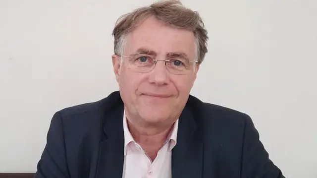 Serges Grouard arongoye igisagara ca Orléans, yambaye ikoti yirabura n'ishati yera. Yambaye kandi n'amarori