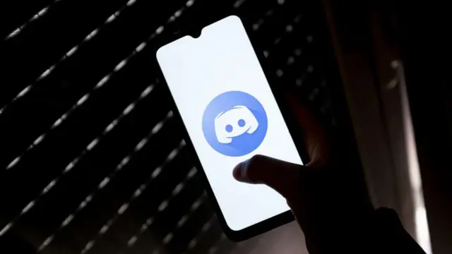 Mão segurando um celular com um logotipo do Discord na tela, em um ambiente escuro 