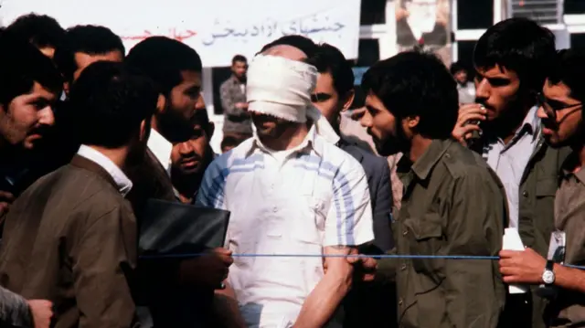 
डोळ्यावर पट्टी बांधलेला ओलीस असलेला अमेरिकन नागरिक आंदोलकांनी वेढलेला आहे. 