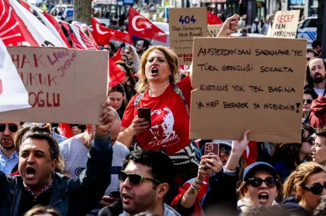 Protesto için toplanan topluluğun içinde iki kişi pankart tutuyor, bir kadın slogan atıyor.
