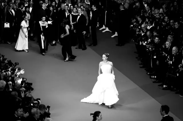 Jennifer Lawrence camina con un vestido blanco por una alfombra roja mientras la fotografían decenas de fotógrafos. La imagen está en blanco y negro.