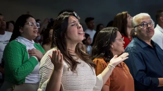 Késia ao lado dos paissite de apostas esportivas brasilculto; ela levanta as palmas da mão para o alto e estásite de apostas esportivas brasilolhos fechados