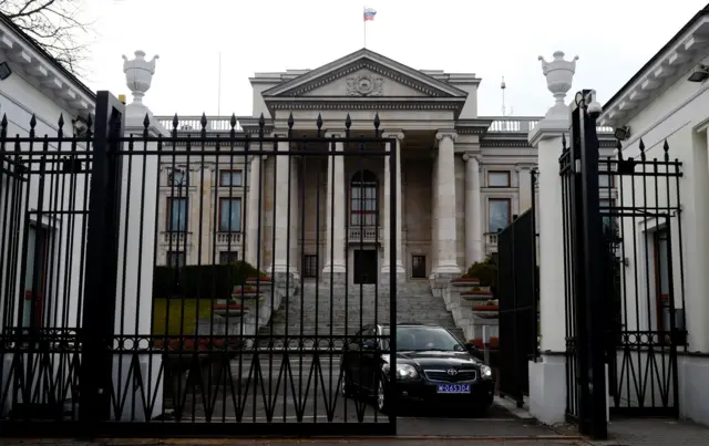 Ambasada Rosji w Warszawie. Duży neoklasyczny budynek, do którego prowadzi ciężka żelazna brama. Na zdjęciu z otwartej bramy wyjeżdża czarny samochód z niebieską tablicą rejestracyjną.