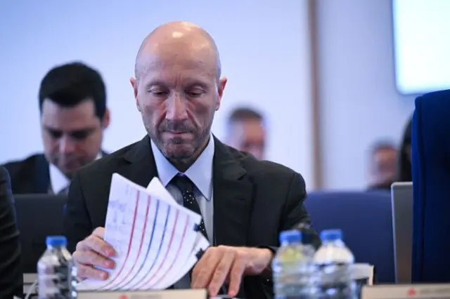 Akçay masasındaki kağıtlar arasında bir şey arıyor gibi görünüyor. Geri planda resmi giyimli birkça kişi daha var, flular. 