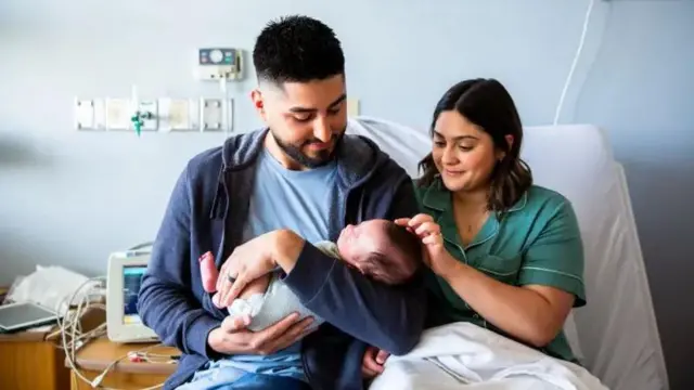 A father and mother sit next to each other in a hospital bed while holding a newborn baby. The father holds the baby in his arms, while the mother strokes its head.