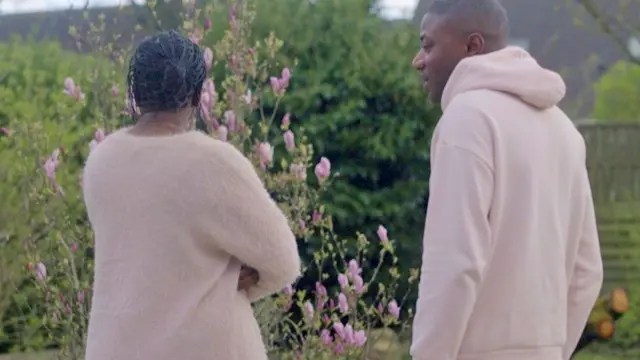 Fatoumata Sylla (à gauche) regarde un jardin fleuri, les bras croisés et le dos tourné à l'appareil photo. Son frère Abdoul (à droite) la regarde - à Paris, en France. Tous deux portent des hauts rose saumon