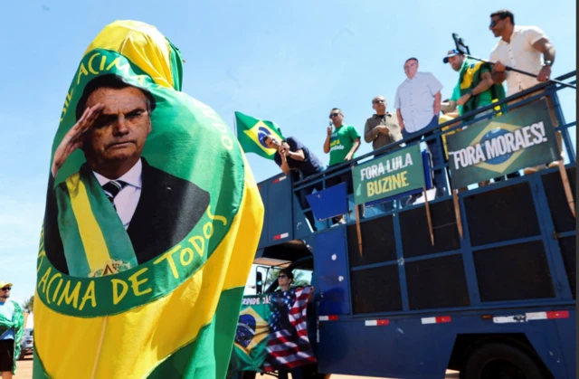 Uma pessoa carrega uma bandeira com a foto do ex-presidente brasileiro Jair Bolsonaro, enquanto apoiadores do ex-presidente se reúnem do lado de fora de seu condomínio, onde ele está em prisão domiciliar, aguardando a última instância de seu julgamento, acusado de planejar um golpe após sua derrota eleitoral em Brasília, Brasil (31 de agosto de 2025).