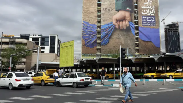 Tahran'da büyük bir caddede zebra geçidinden geçen bir adam ve akan trafik. Arka planda bir reklam panosunda Tahran yönetiminin bir propaganda posterinde Hürmüz Boğazı şeklinde resmedilen mavi bir kumaş parçasını sıkan bir yumruk görülüyor.