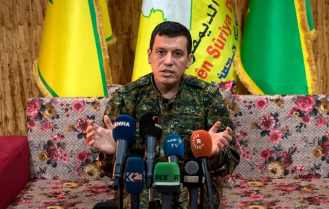 مظلوم عبدي جالسا أمام كاميرات وميكروفونات الأعلام وفي الخلفية العلم الأصفر