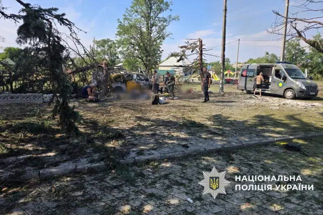 Спалені дерева, плитка, засипана землею. Попредеу видно людей, які стоять біля руїн машини після обстрілу