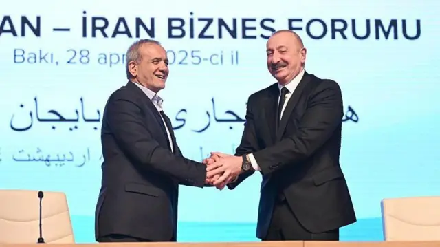 Двое мужчин в темных костюмах, улыбаясь, жму друг другу руки на фоне экрана с рекламой бизнес-форума в Баку