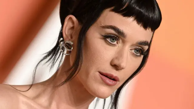 Katy Perry está maquiada e com cabelos presos. Ela usa brincos pratas, franja.