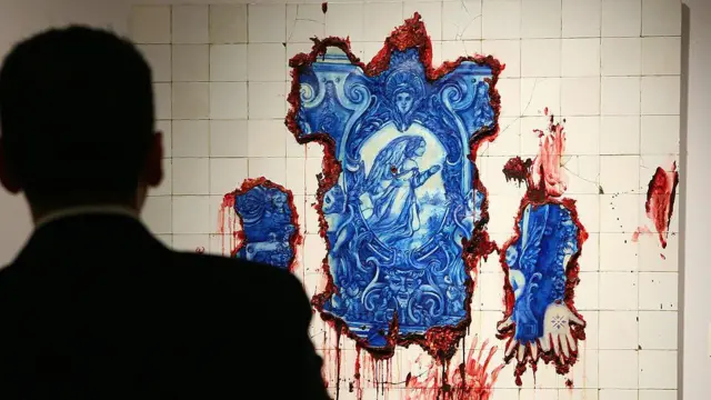 A silhueta de um homem observa uma obra formada por azulejos azuis de inspiração portuguesa rasgados por aberturas que revelam uma massa vermelha semelhante a carne e sangue.