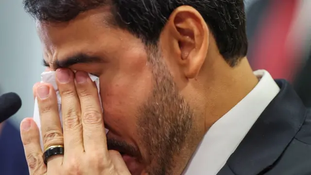 Jorge Messias, de terno, aparece em close, com os olhos fechados e o rosto parcialmente coberto pelas mãos enquanto segura um lenço. Ao lado, há um microfone.