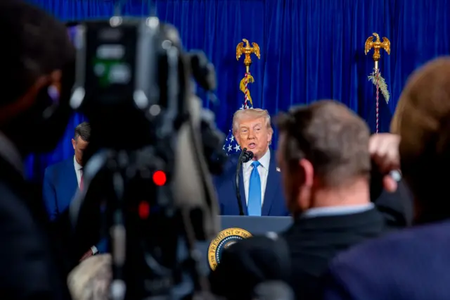 Trump de fondo. Delante, una cámara de televisión y varias personas.