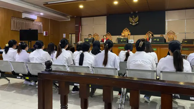 Dua baris orang berpakaian putih duduk di bangku di ruang pengadilan, menghadap panel hakim
