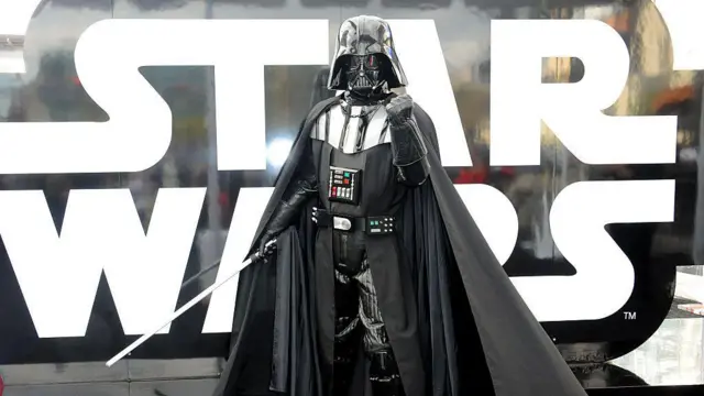 imagen de una persona con un traje de darth vader. tiene el puño cerrado y levantado hacia el frente y en la otra mano sostiene un sable de luz. 