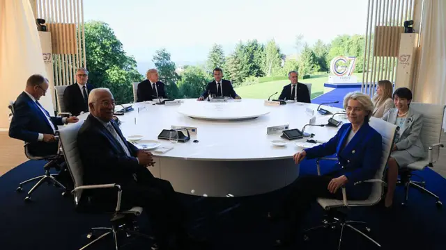Patrząc od lewej: António Costa, przewodniczący Rady Europejskiej, kanclerz Niemiec Friedrich Merz, premier Wielkiej Brytanii Keir Starmer, prezydent USA Donald Trump, prezydent Francji Emmanuel Macron, premier Kanady Mark Carney, premier Włoch Giorgia Meloni, premier Japonii Sanae Takaichi oraz przewodnicząca Komisji Europejskiej Ursula von der Leyen siedzą wokół dużego, okrągłego białego stołu. W tle, za dużym oknem, widoczny jest soczyście zielony krajobraz oraz logo G7.
