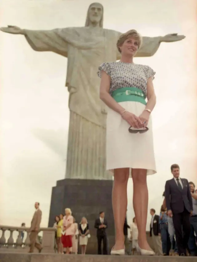 A princesa usa uma saia branca e uma blusa estampada, com um cinco verde. No fundo da foto há várias pessoas.