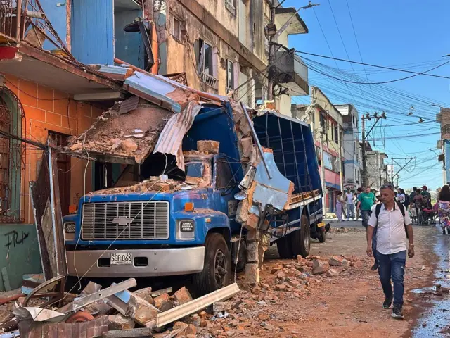 10 Ağustos 2026 tarihinde Kolombiya’da meydana gelen depremde bir binanın yıkıntıları mavi bir kamyonun üzerinde ve sokaktan insanlar geçiyor