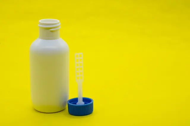 Una botellita blanca destapada. Al lado la tapa, conectada a una pequeña espátula plástica.