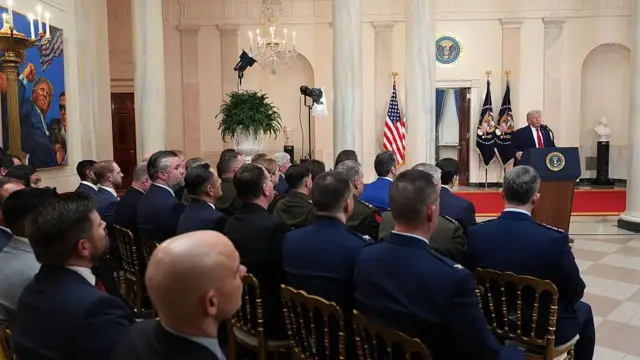 Trump'ın konuşma yaptığı geniş salonda çok sayıda yetkili onu dinliyor. Hepsini arkadan görüyoruz.