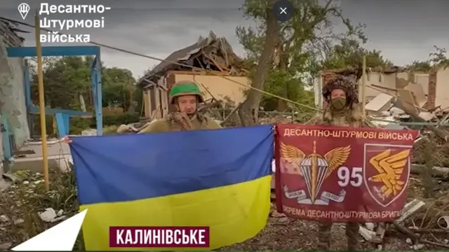 Двоє військових ЗСУ розгорнули і тримають прапори України і 95 бригади ДШВ на фоні зруйнованих будинків