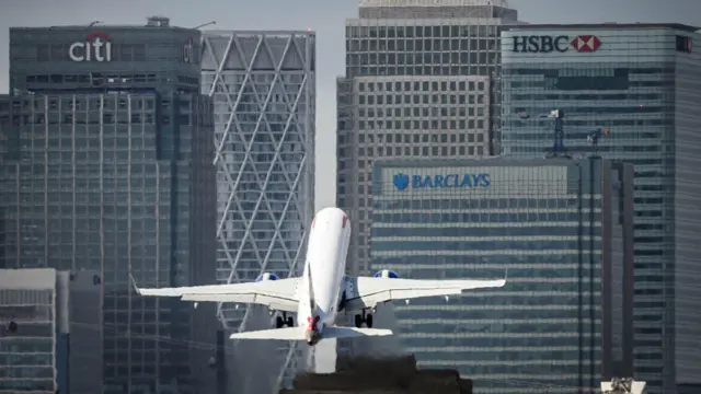 Um avião decola diante de prédios de escritórios com fachada de vidro em Canary Wharf, em Londres, com logotipos visíveis do Citi, HSBC e Barclays