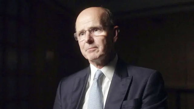 杰瑞米·弗莱明爵士身穿深海军蓝色西装外套,肩部线条分明,搭配洁白衬衫与淡蓝色纹理领带,坐在一个光线昏暗的室内环境中,背景呈深色中性色调并带有柔和阴影。灯光突显西装布料与领带质感,营造正式专业氛围。