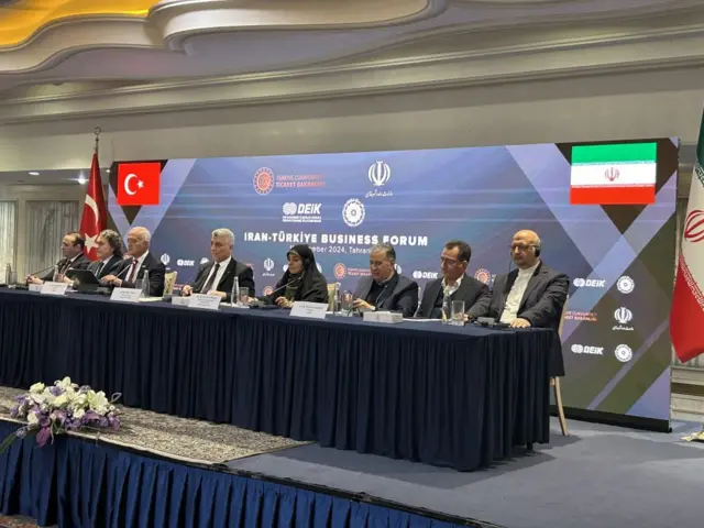 Ömer Bolat bir masada, İranlı yetkililerle oturuyor. Arkada Türkiye ve İran bayrakları var.