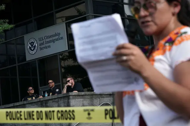 USCIS ရုံးရှေ့မှာ ရပ်နေတဲ့ အမျိုးသမီးတစ်ဦးနဲ့ အနောက်ကနေထိုင်ကြည့်နေတဲ့ရဲ ၃ဦး 