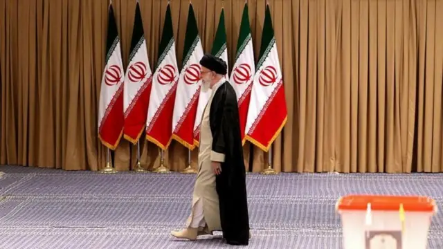 আয়াতুল্লাহ আলী খামেনি, পেছনে ইরানের সাতটি পতাকা। সামনে একটি ভোটের বাক্স।