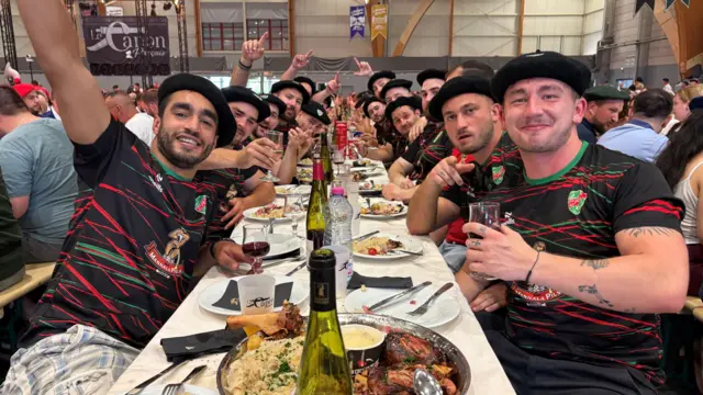 Un grupo de jóvenes vestidos con camisetas deportivas a juego y boinas negras está sentado a una larga mesa con fuentes de comida y vino.