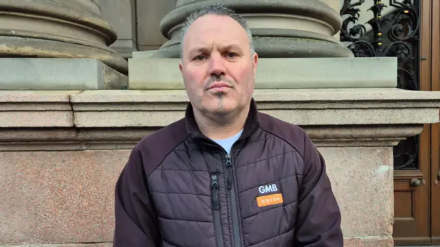 Chris Mitchell, un hombre con el pelo corto canoso, bigote y perilla, de pie afuera, mirando directamente a la cámara. Lleva una chaqueta marrón con cremallera y un pequeño logotipo de GMB UNION.