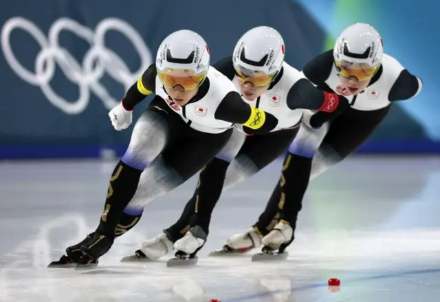 白黒のウエア、白のヘルメットを着けた3人が縦に並んでカーブを滑っている