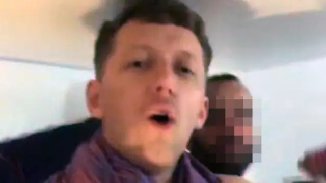 Скріншот із відео, на якому чоловік говорить у камеру, а позаду нього стоїть інший чоловік із розмитим обличчям.