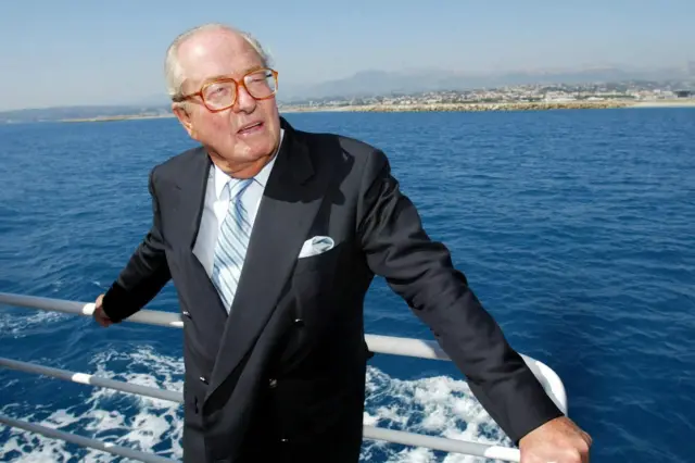 Jean-Marie Le Pen wearing a dark navy suit with a light blue tie and white pocket square stands on the deck of a boat, holding onto the railing. The boat is on a calm blue sea.
