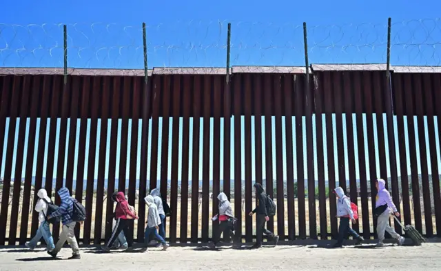 migrantes caminan frente a una verja en la frontera entre ee.uu. y méxico
