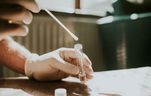 A hand wearing a latex glove holds a small clear tube and dips a cotton swab into the liquid.