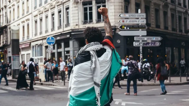 مردی معترض در خیابان با پرچم فلسطین و چفیه دستش را مشت کرده